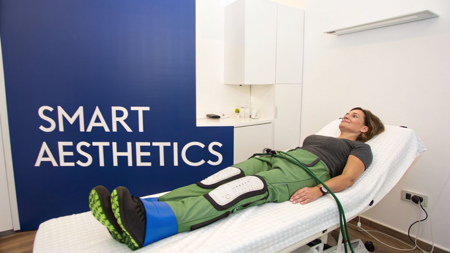 A smiling woman lies on a bed, undergoing a leg treatment with specialized green and blue compression pants, in a modern aesthetics clinic.