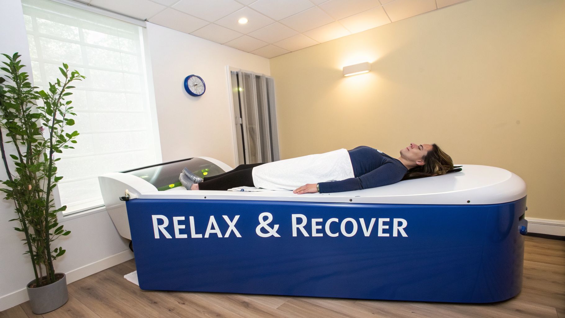 A woman lies on a blue and white 'RELAX & RECOVER' therapy machine in a bright room.